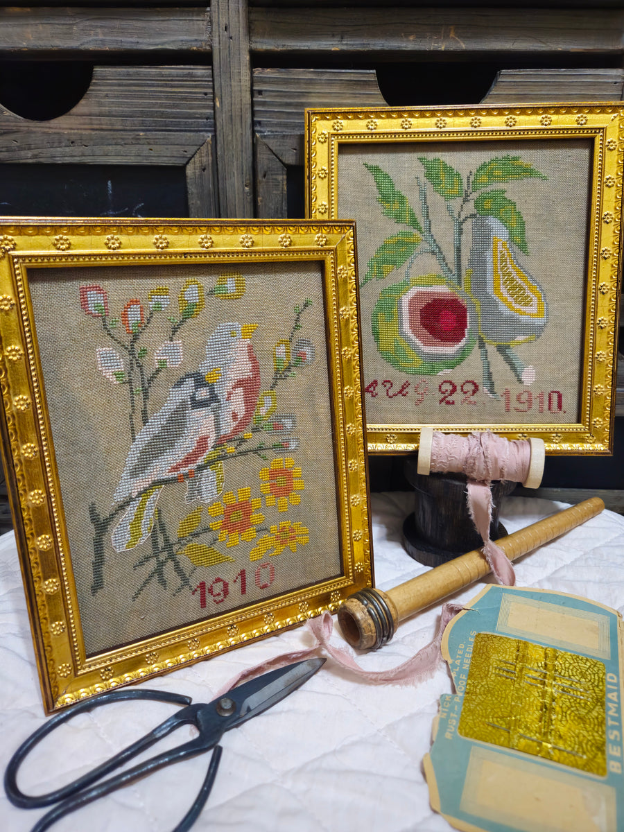 Two framed needlepoint pieces with gold frames, a pair of scissors, and a spool of thread on a wooden surface.