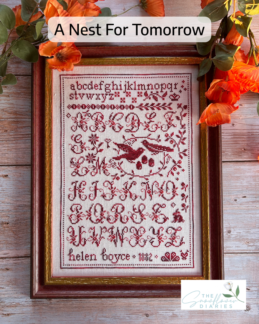 A framed cross-stitch sampler with a bird and floral design, titled 'A Nest For Tomorrow,' displayed on a rustic wooden surface with orange flowers around it. The sampler reads 'helen boyce 1892' at the bottom, and a 'The Sewing Diaries' logo appears in the lower right corner.