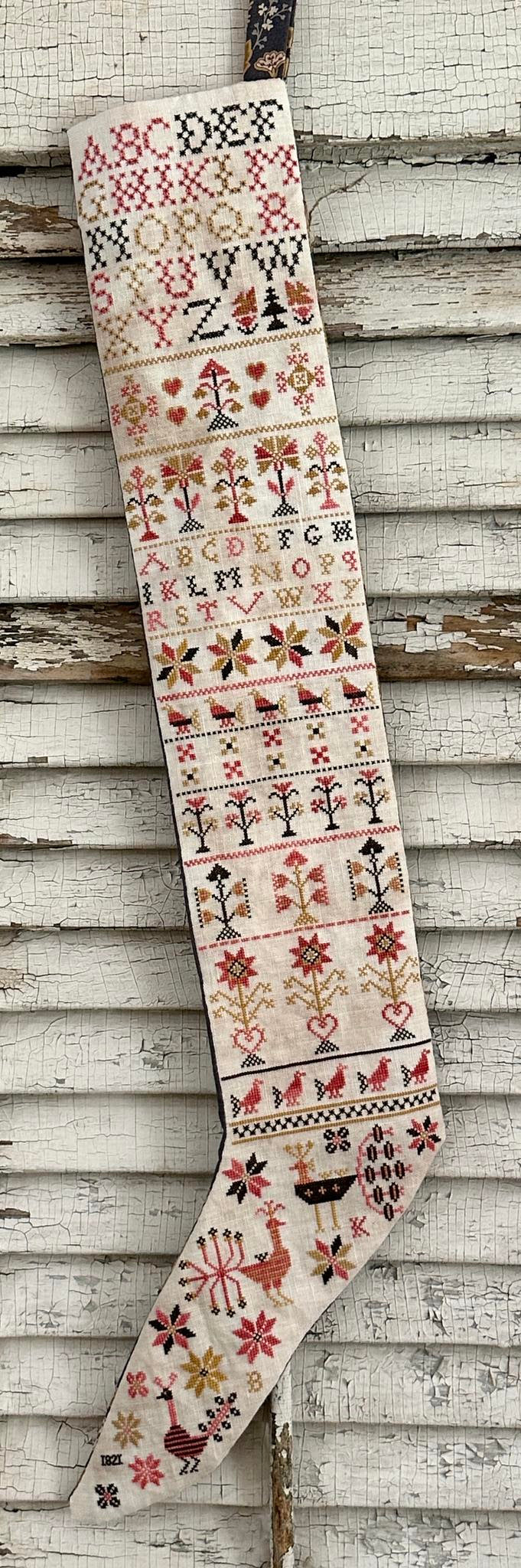 A vintage-style Christmas stocking with intricate embroidered patterns, hanging against a rustic white-painted wooden wall.