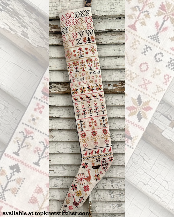 A Christmas stocking with intricate multicolor embroidery, hanging against a rustic white-painted wooden wall. The stocking features various festive patterns including snowflakes, trees, birds, and letters.