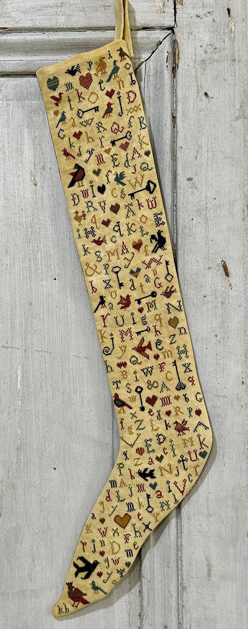 A tall, narrow beige stocking or hanging textile decorated with dense embroidered-style lettering and small motifs in red, blue, black, and brown, shown against a weathered white wooden background.
