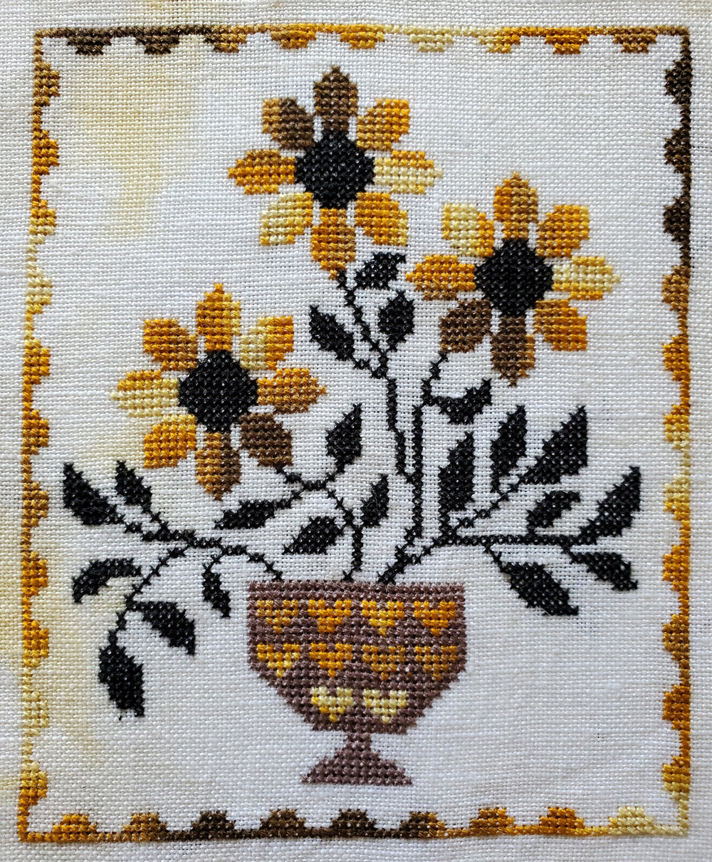 Cross-stitched design of sunflowers in a vase on white fabric.