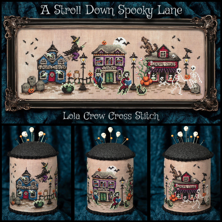 A framed cross-stitch artwork titled "A Stroll Down Spooky Lane" with three smaller views below showing the stitched fabric containers. The design depicts Halloween-themed houses, bats, pumpkins, a witch, skeletons, and streetlamps on a beige fabric background, set against a dark blue textured backdrop.