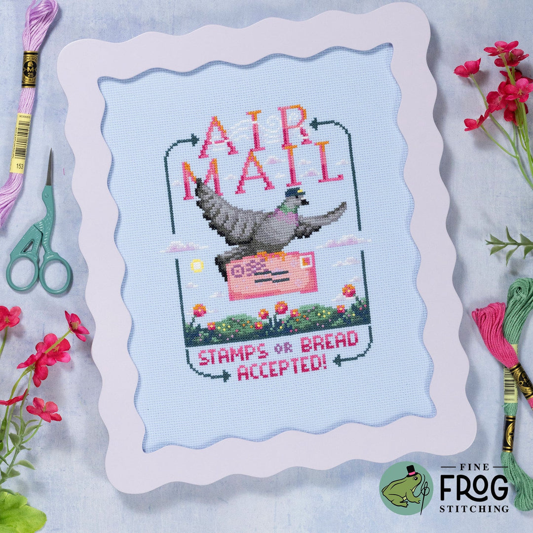 A framed cross-stitch design featuring a pigeon carrying a pink mail envelope with the words “AIR MAIL” and “STAMPS OR BREAD ACCEPTED!” on a light blue fabric background. The frame is shown on a styled flat-lay with embroidery floss, scissors, and flowers around it.