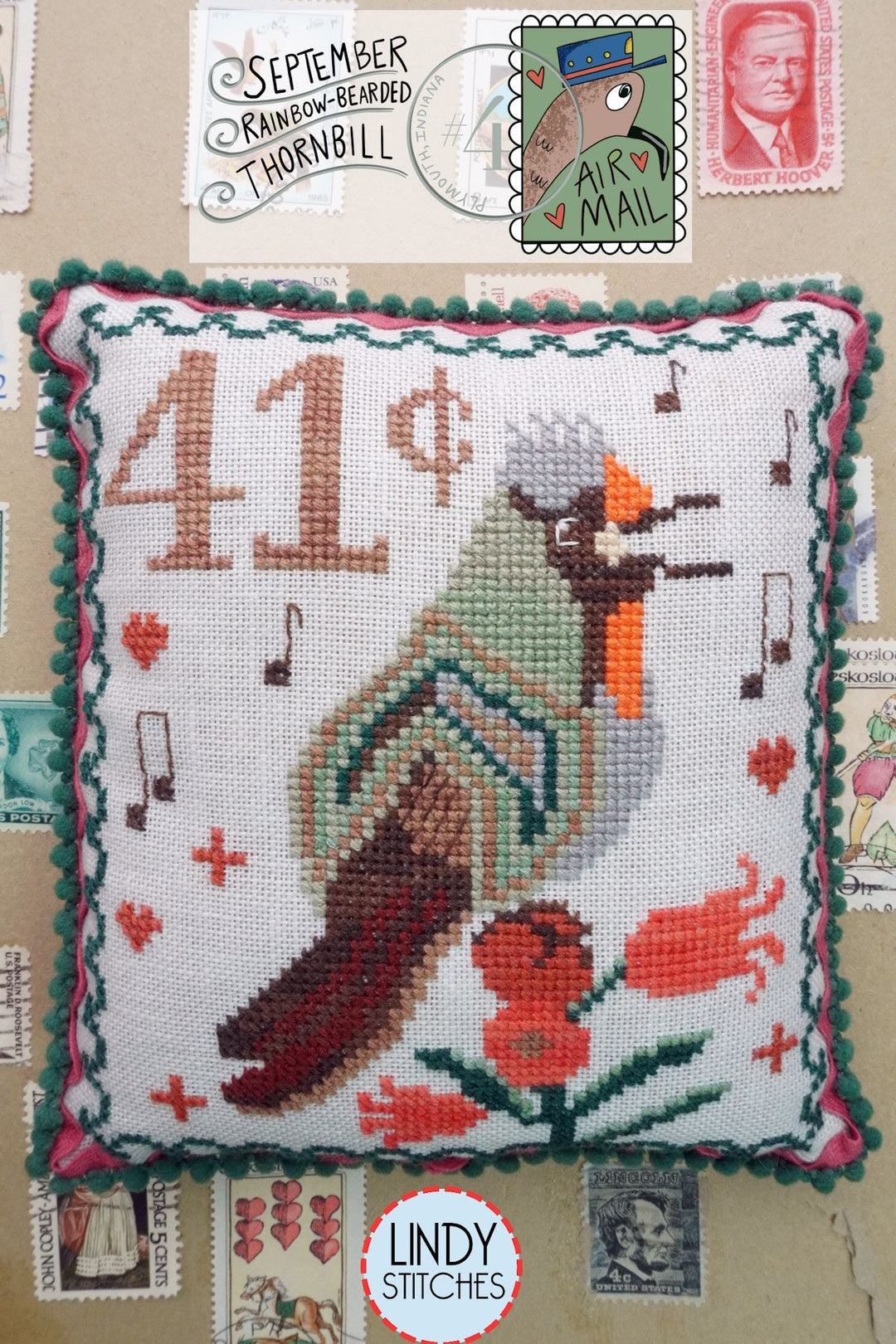 Cross-stitched pillow with a bird design and Lindy Stitches branding, surrounded by vintage stamps.