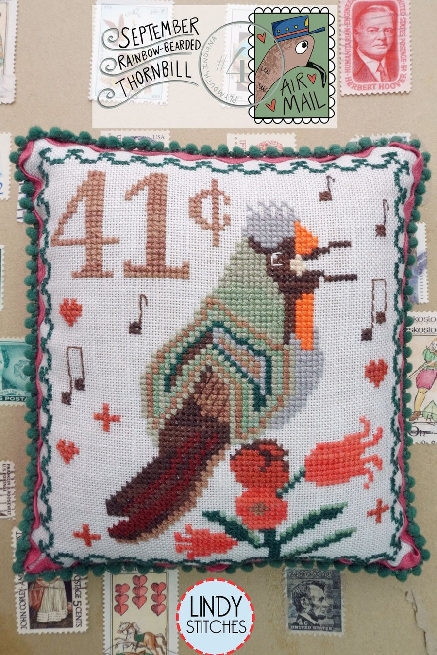 Cross-stitched pillow with a bird design and Lindy Stitches branding, surrounded by vintage stamps.