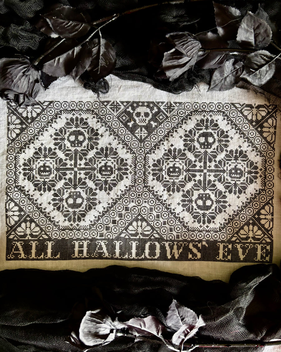 Decorative cloth with skull pattern and 'All Hallows' Eve' text, surrounded by dark leaves.