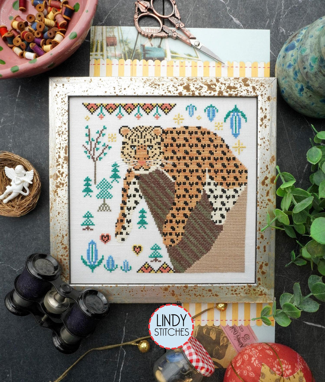 a cross-stitch kit depicting an Amur leopard displayed alongside crafting tools like scissors and a spool of thread,
