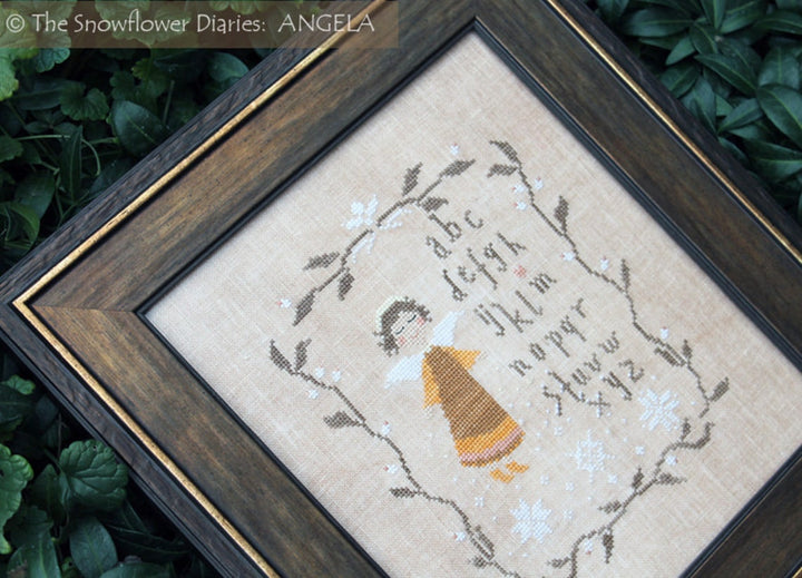 A framed cross-stitch sampler featuring a child in a yellow dress surrounded by white flowers and alphabet text, set in a dark wood frame with green foliage in the background.