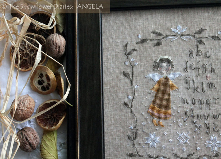 A framed cross-stitch artwork on linen fabric showing a small angel figure, floral border, and alphabet text. The frame is dark wood, and dried orange slices, nuts, and straw are arranged beside it as styling props.