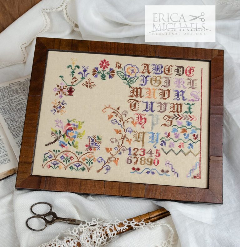 Framed cross-stitched sampler with alphabets, numbers, and floral patterns on a white fabric background.