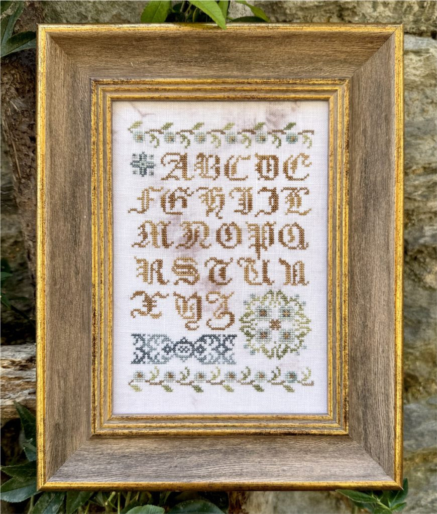 A framed cross-stitch sampler featuring an alphabet and decorative floral motifs, set in a rustic wooden frame with a gold-toned inner border, photographed against a natural stone and green foliage background.