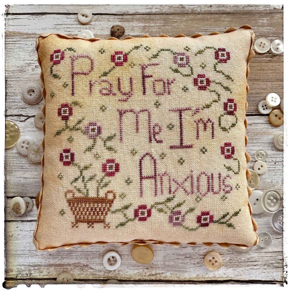 Decorative pillow with cross-stitched text and flowers on a wooden background with buttons.