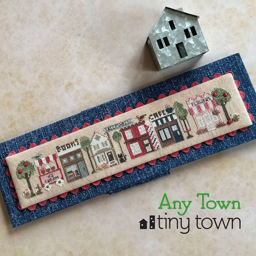 Cross-stitched design of a town with 'Any Town tiny town' branding on a textured surface.