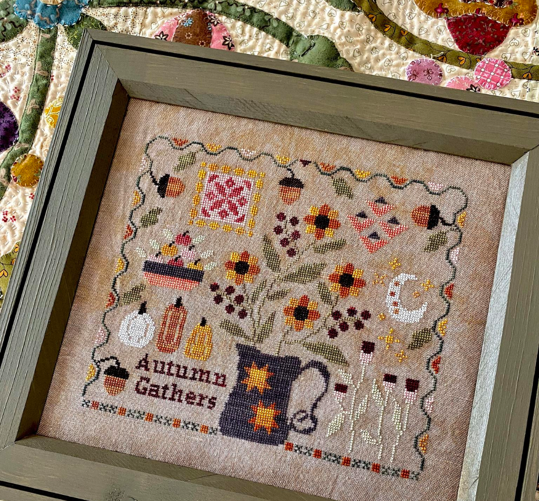 Framed cross-stitched piece with autumn-themed design on a textured fabric background.