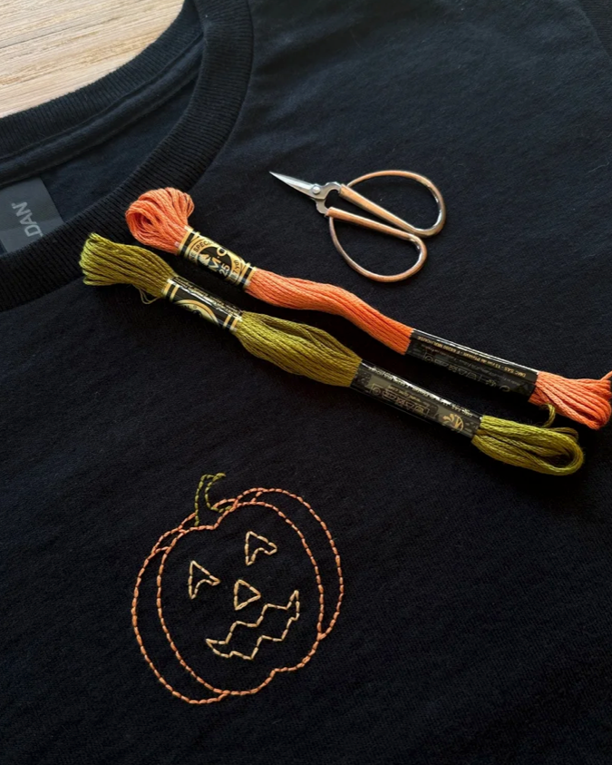 A close-up of a black t-shirt featuring an embroidered orange jack-o'-lantern, styled with embroidery floss in orange and olive green, and small embroidery scissors on a light wood background.