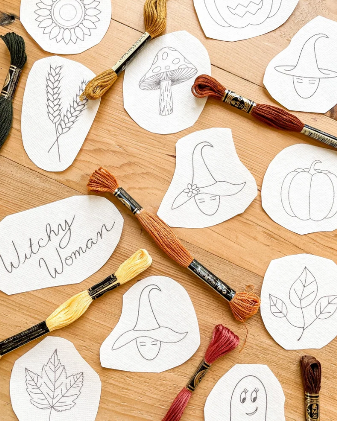 A styled flat-lay of embroidery floss spools and embroidered white fabric patches with autumn-themed line drawings, including a witch's hat, pumpkin, mushroom, wheat, leaves, and a ghost, arranged on a light wood surface.