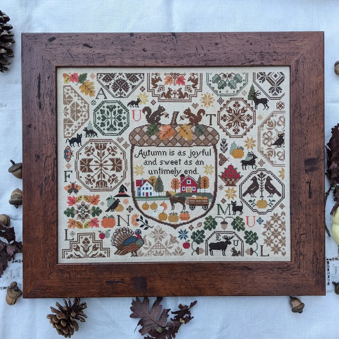 Framed cross-stitched sampler with autumn-themed designs on a white background.