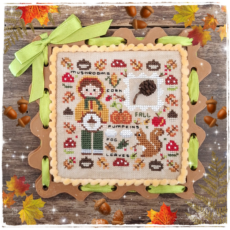 A finished cross-stitch sampler featuring an autumn theme, displayed on a scalloped wooden frame with green ribbon accents. The sampler depicts a girl, pumpkins, mushrooms, corn, and fall elements on a beige fabric background, set against a rustic wood backdrop with scattered acorns and autumn leaves.