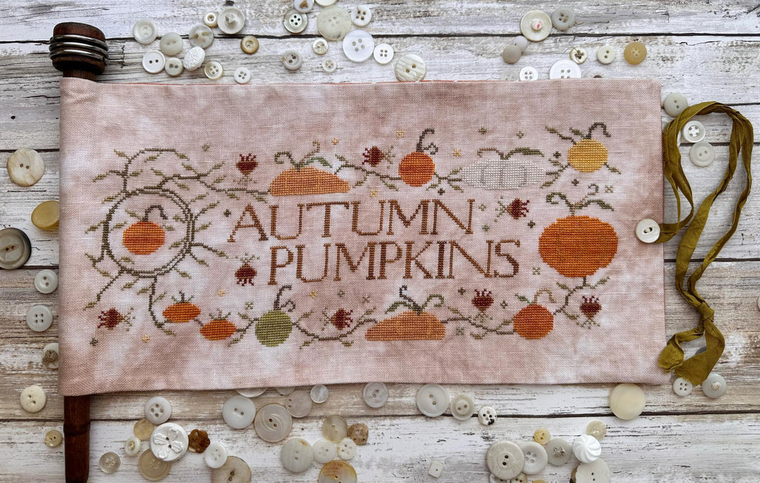 A square fabric cross-stitch sampler with 'AUTUMN PUMPKINS' stitched in the center, surrounded by embroidered pumpkins, acorns, and autumn leaves. The sampler is laid flat on a rustic white-washed wooden surface, surrounded by scattered decorative buttons and a green ribbon.