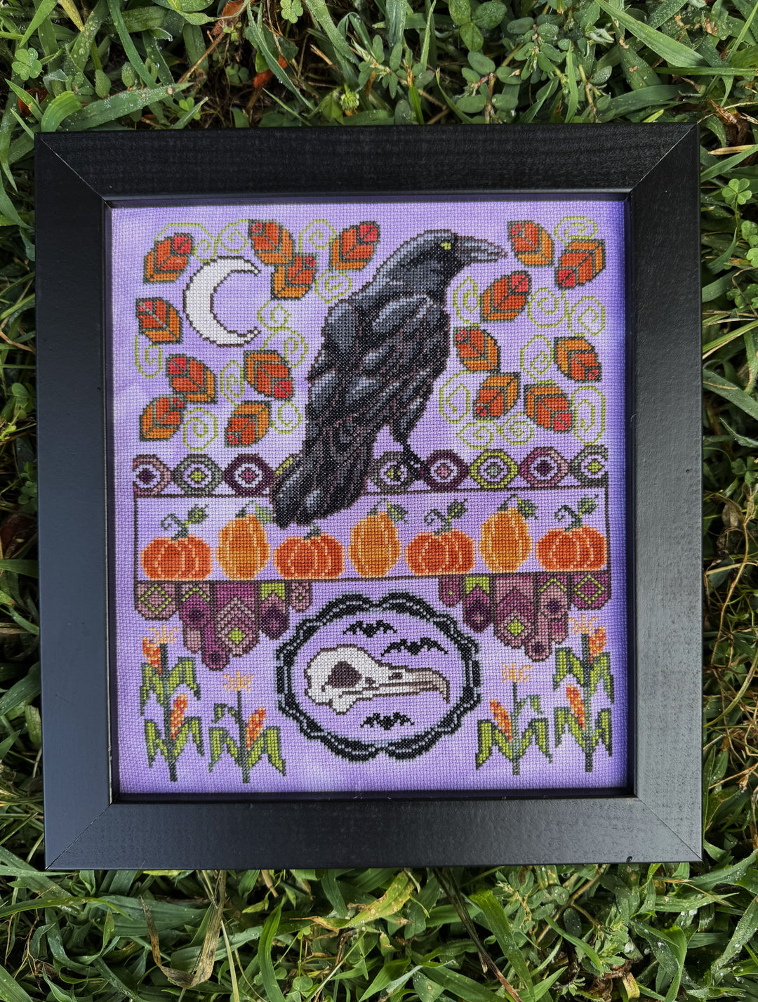 A framed cross-stitch artwork featuring a black raven, orange pumpkins, autumn leaves, a crescent moon, and a raven skull motif on a lavender background, placed on green grass.