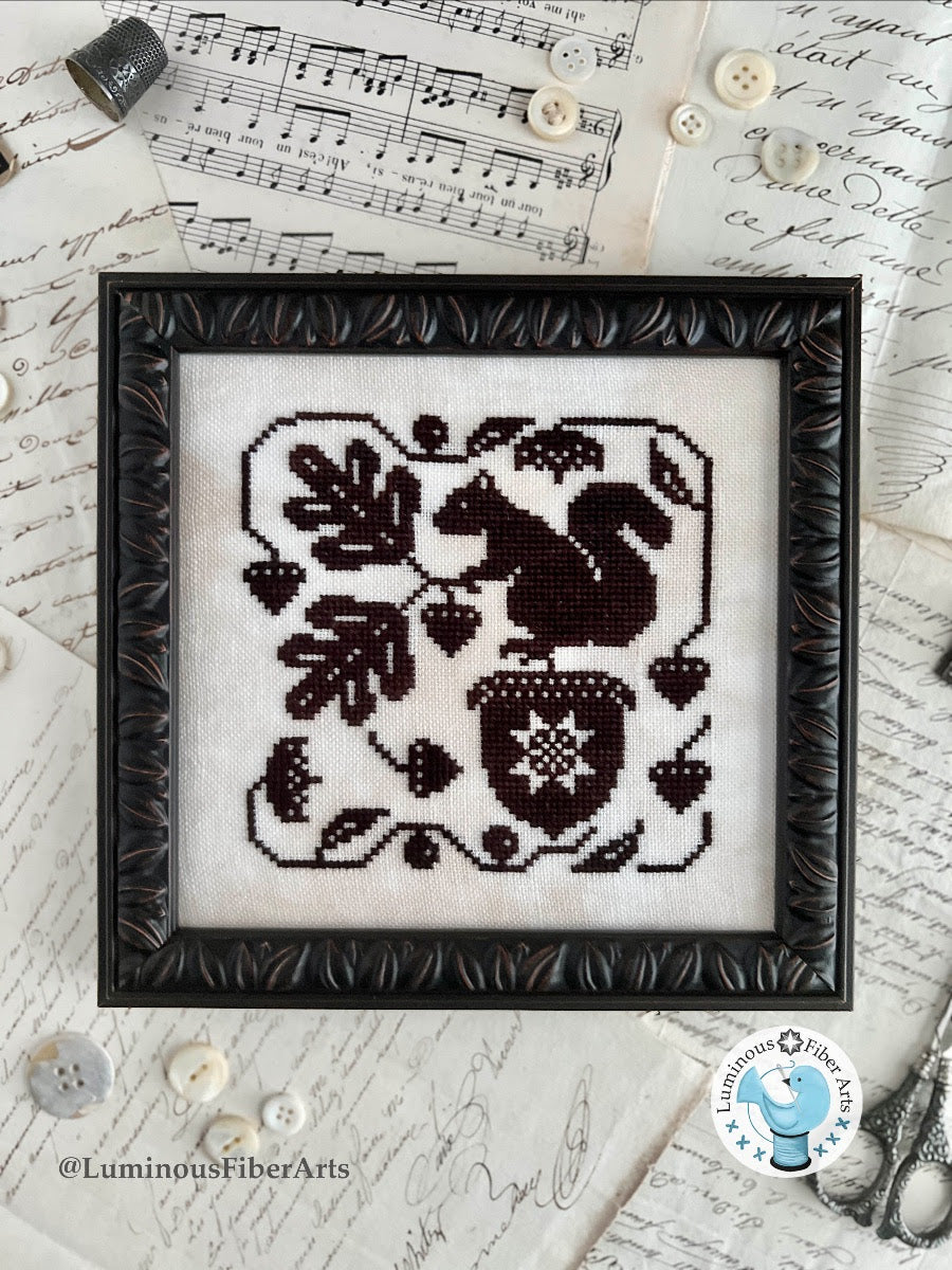 Framed cross-stitched pattern of a squirrel with acorns on a textured surface with music sheets and buttons.