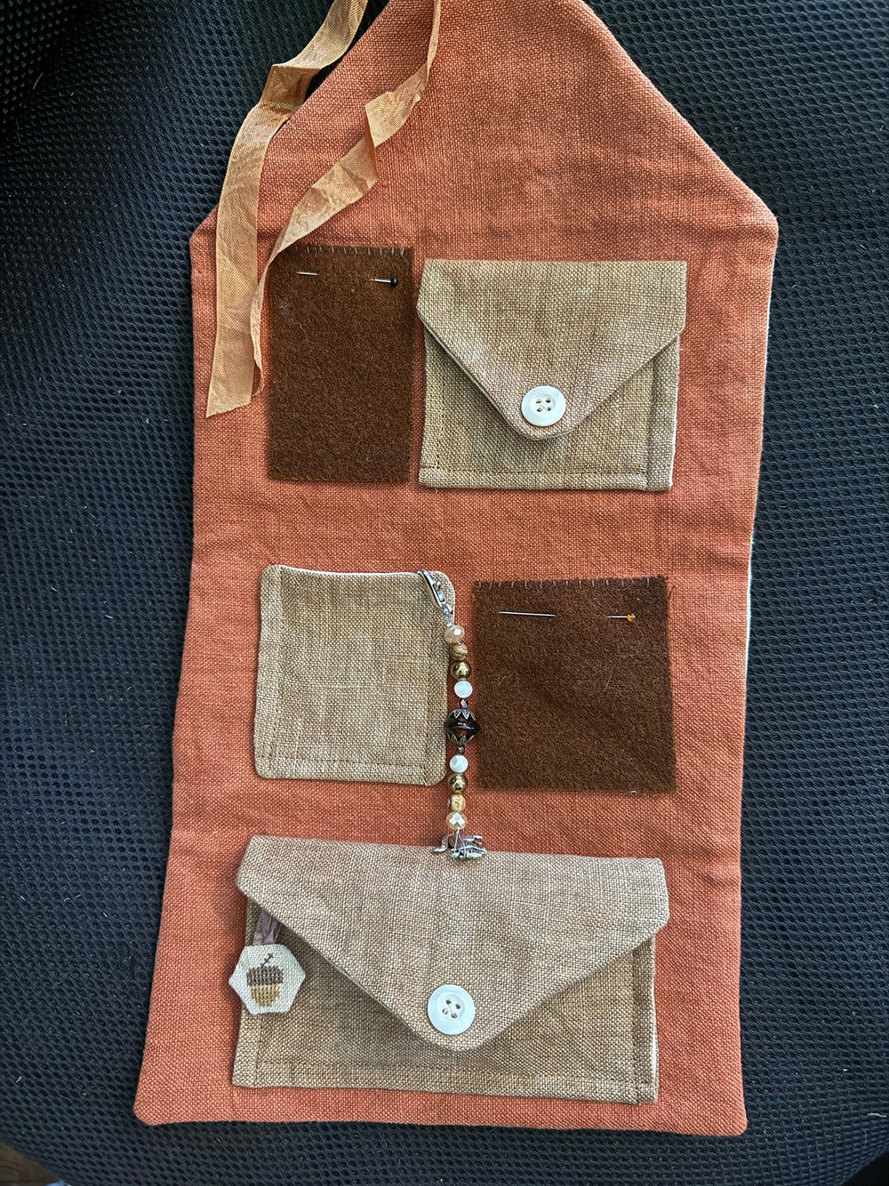 A rustic fabric organizer pouch with multiple pockets in terracotta, tan, and dark brown, featuring a beaded charm and a small wooden tag, laid flat on a black textured background.