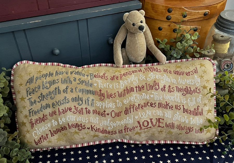 Decorative pillow with text and a teddy bear on a table with plants and a pot.