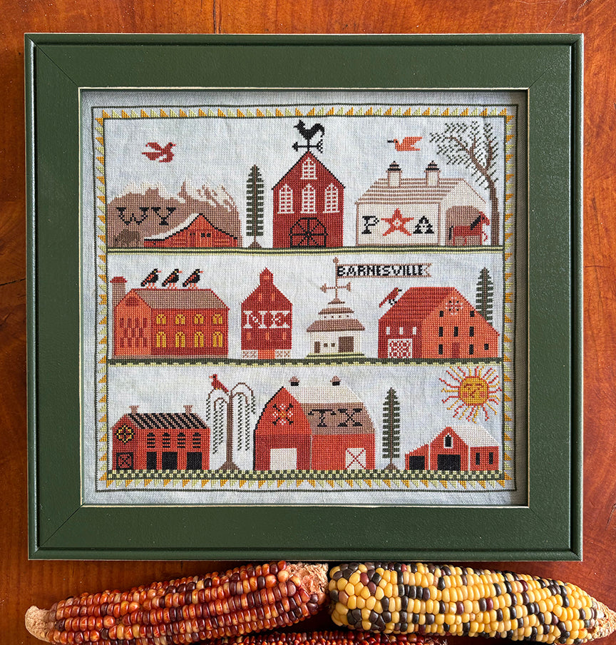 Framed cross-stitched pattern of barns and village buildings on a wooden surface with corn ears.