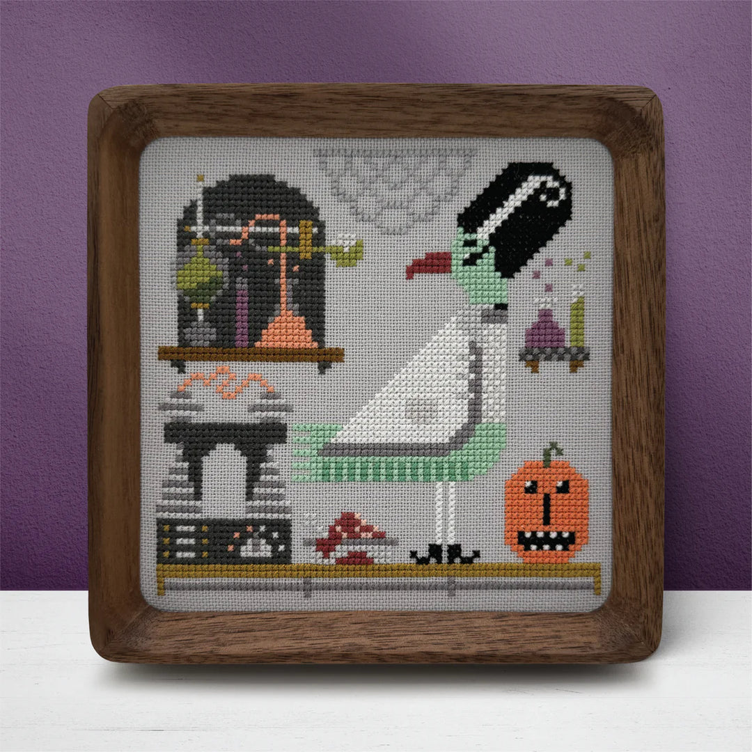 Framed cross-stitched artwork of a seagull and a cat with Halloween-themed elements on a purple background.