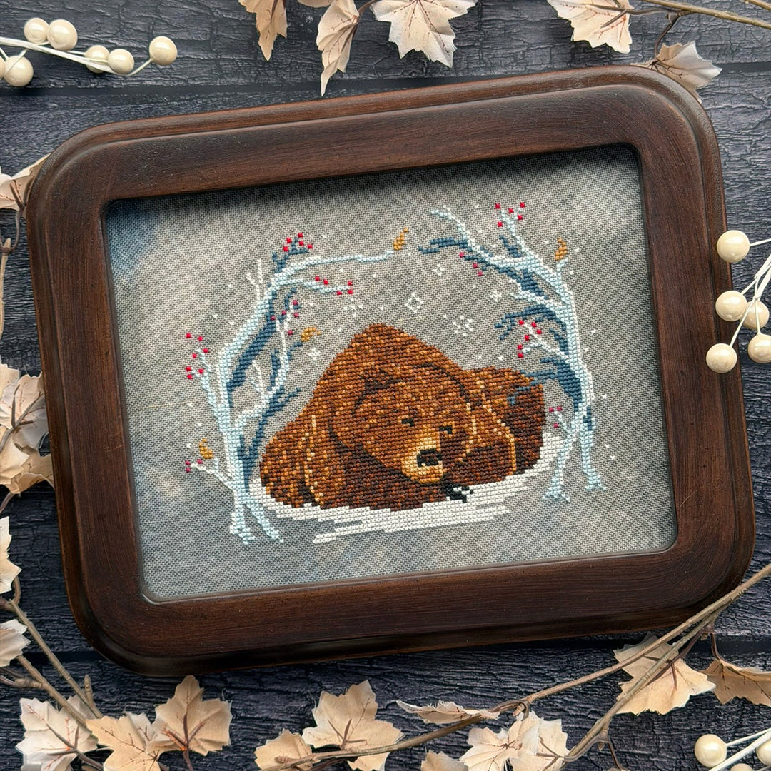 A framed cross-stitch artwork showing a brown bear in a snowy winter scene, surrounded by decorative leaves and small white beads on a dark wooden surface.