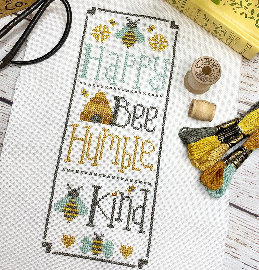 Cross-stitched text with bee designs on fabric, surrounded by embroidery threads and tools.