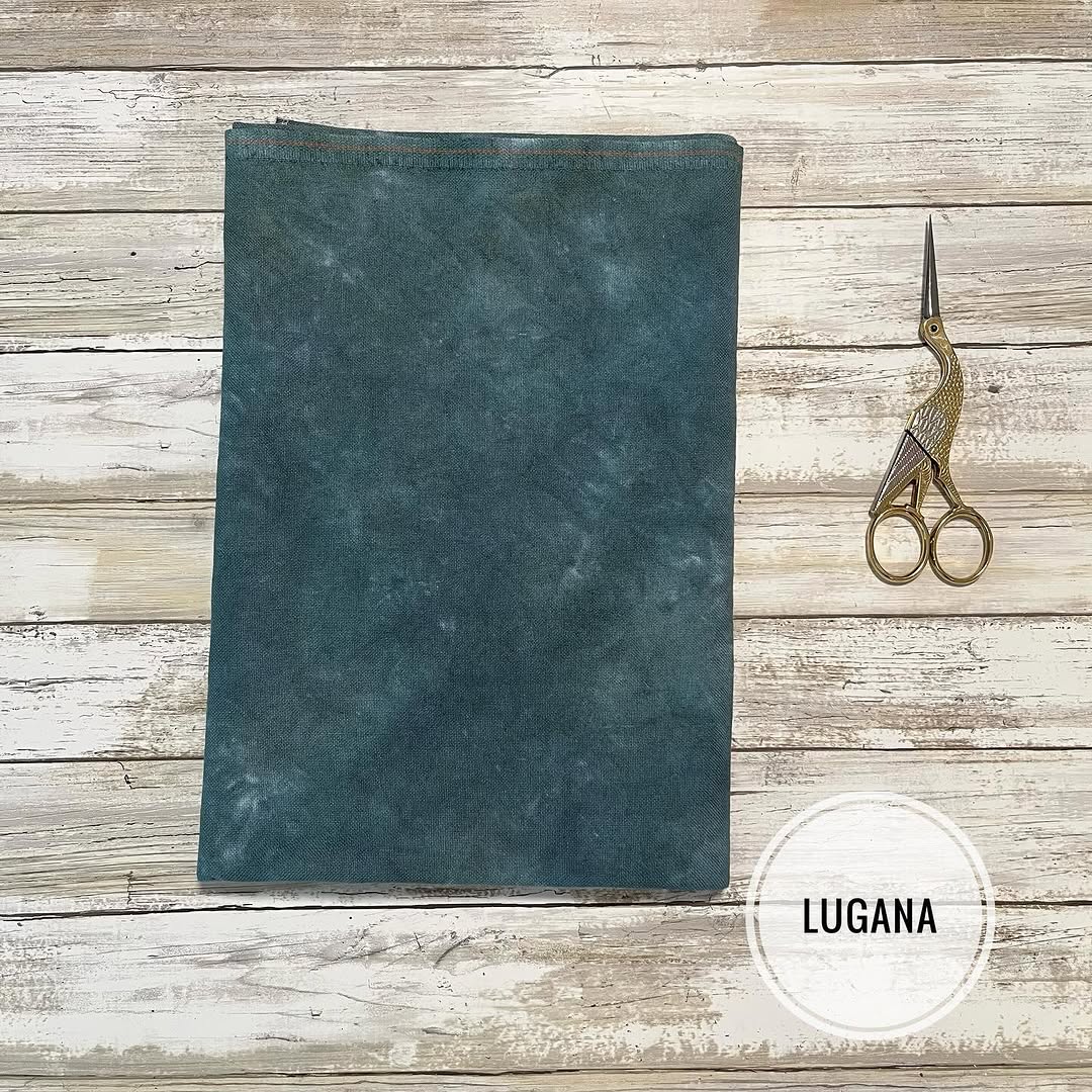 Teal fabric sample with gold scissors on a wooden surface, featuring the brand 'Lugana'.