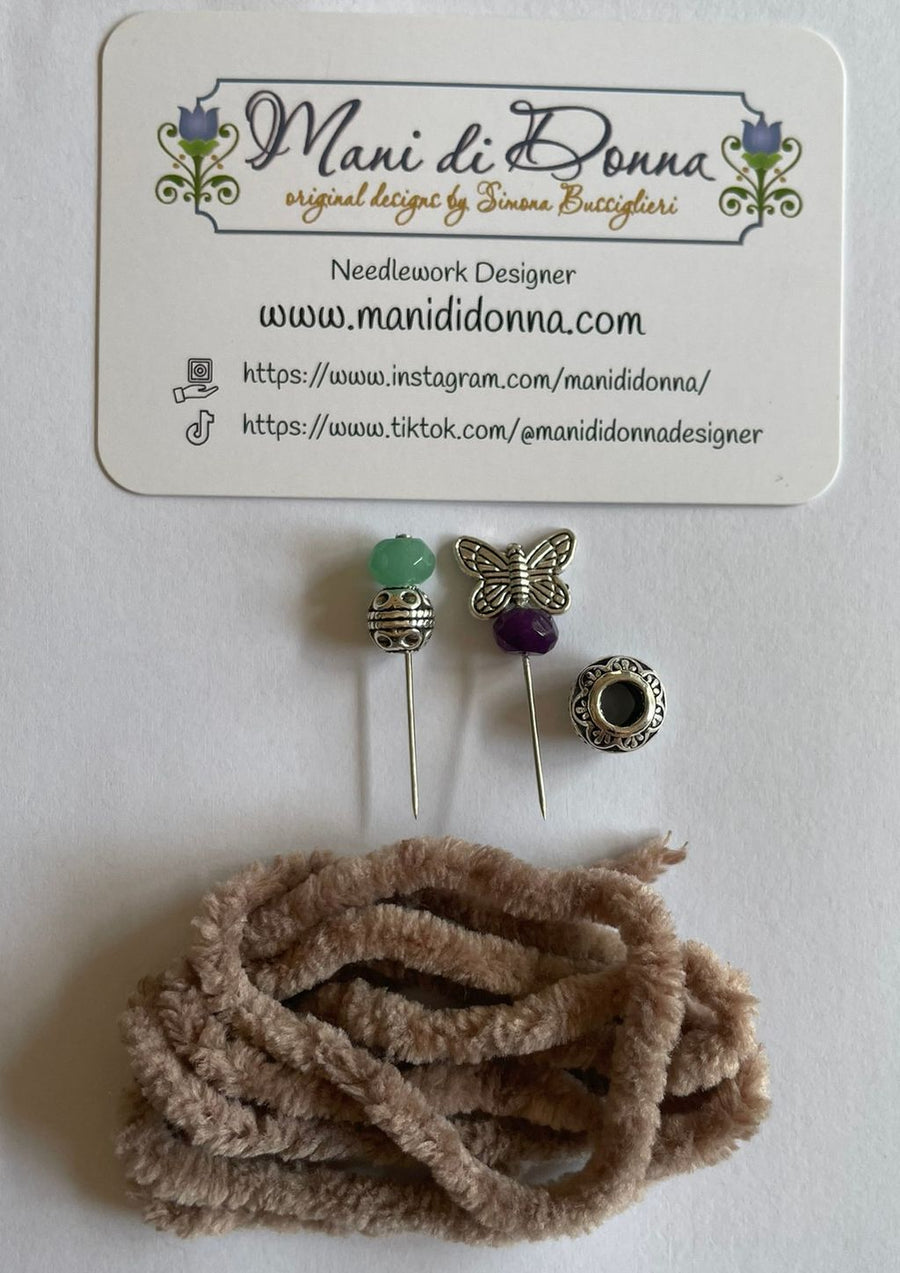 A needlework design set by Mani di Donna, featuring three decorative pins (green bead, butterfly, purple bead, and a round bezel), a coiled bundle of tan chenille stem cord, and a branded white card.