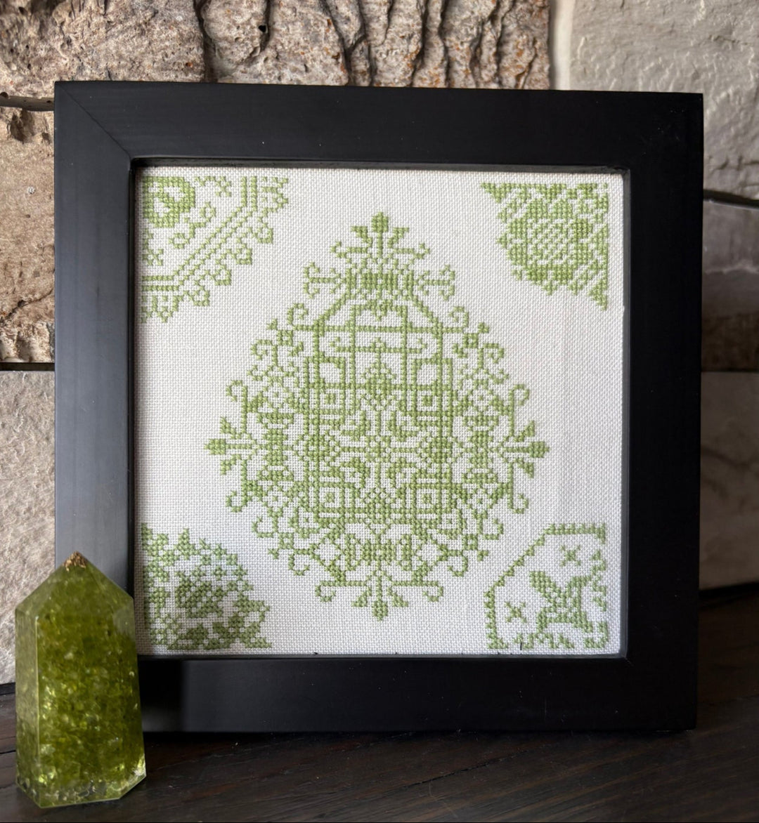 Framed cross-stitched pattern with green design on a white background, leaning against a stone wall.