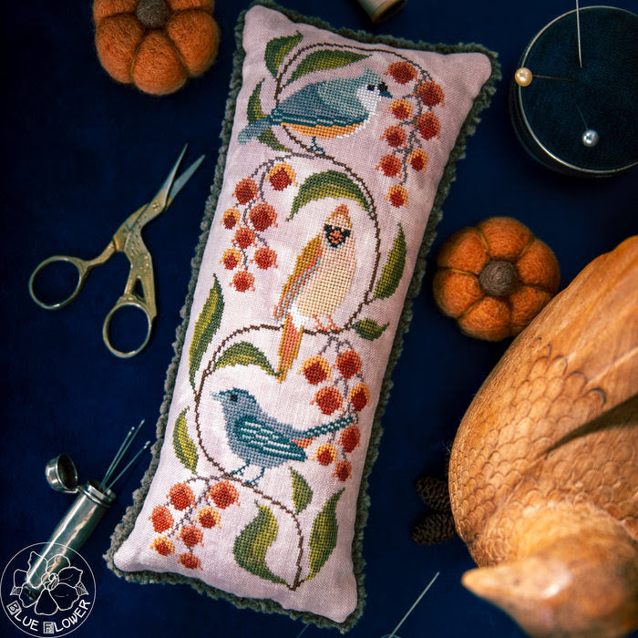 A rectangular embroidered pillow with a pink fabric base, featuring two blue birds and orange berries stitched on a dark navy background, surrounded by autumn-themed props including pumpkins, scissors, and a wooden ornament.