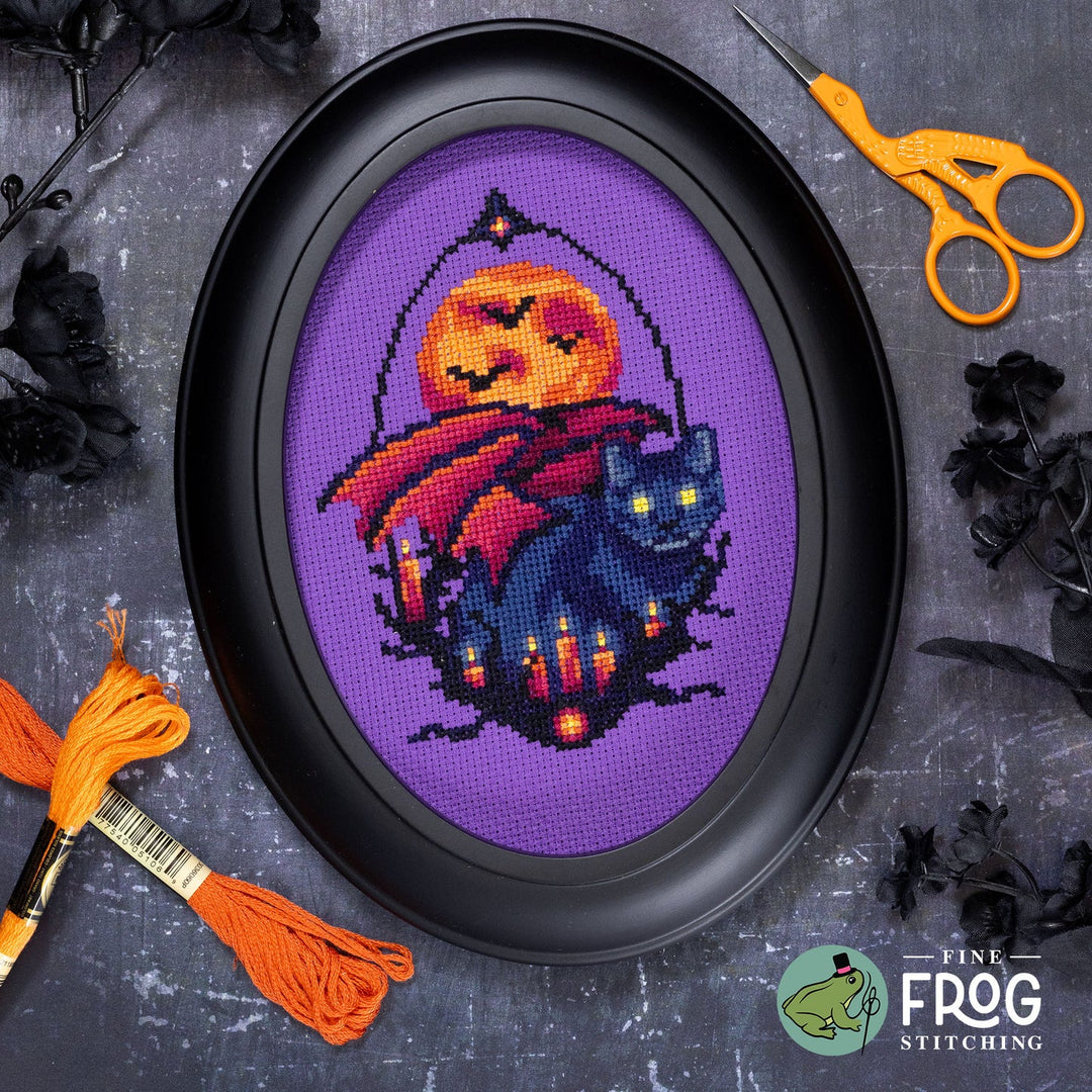 A framed cross-stitch artwork featuring a purple background with a glowing orange moon, a red cape, and a black cat among candles. The oval piece is displayed on a dark textured surface with craft props including orange embroidery floss, orange-handled scissors, and black decorative leaves.