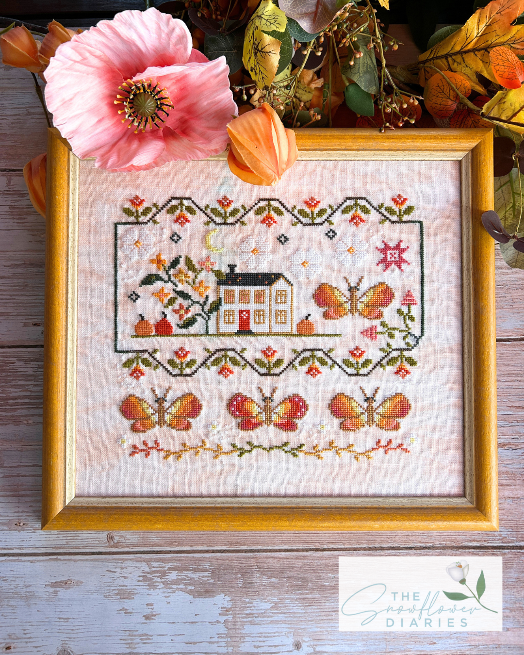 A framed cross-stitch artwork featuring a small house, flowers, and butterflies on a light fabric background, styled with autumn leaves and a pink flower on a rustic wooden surface. A 'The Needlework Diaries' logo appears in the lower right corner.