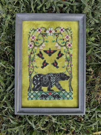 A framed cross-stitch artwork featuring a black bear and floral vine motifs on a yellow-green fabric background, placed on green grass.