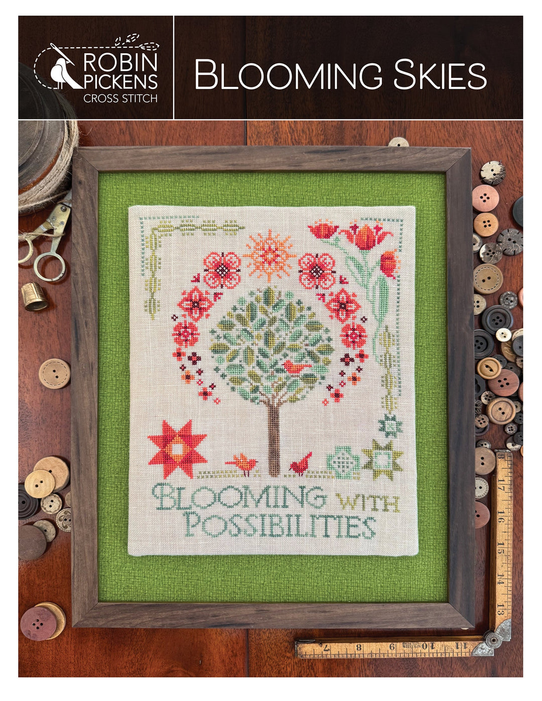 Cross-stitch pattern with floral design and text, framed on a wooden background with sewing tools.