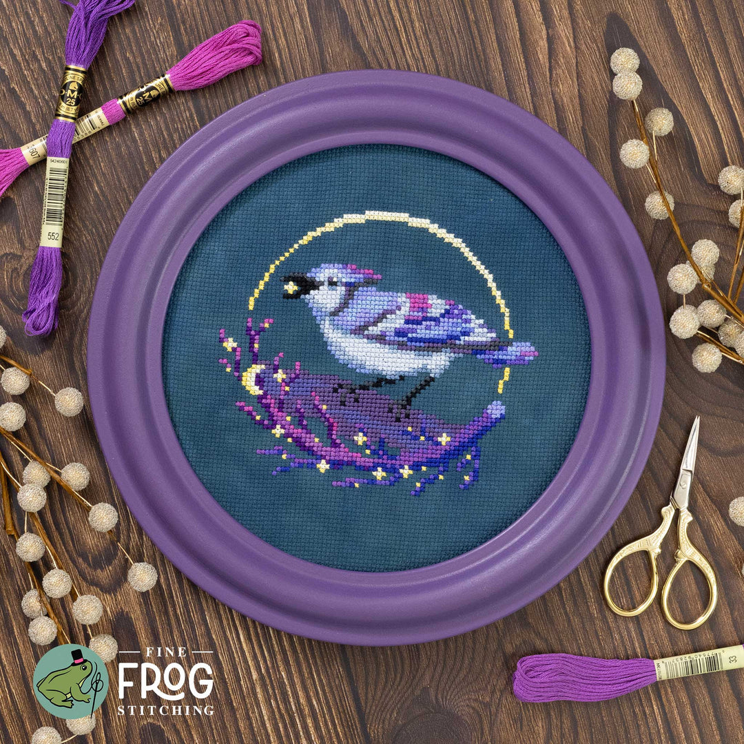 Embroidery hoop with a bird design on a wooden surface with embroidery threads and scissors.