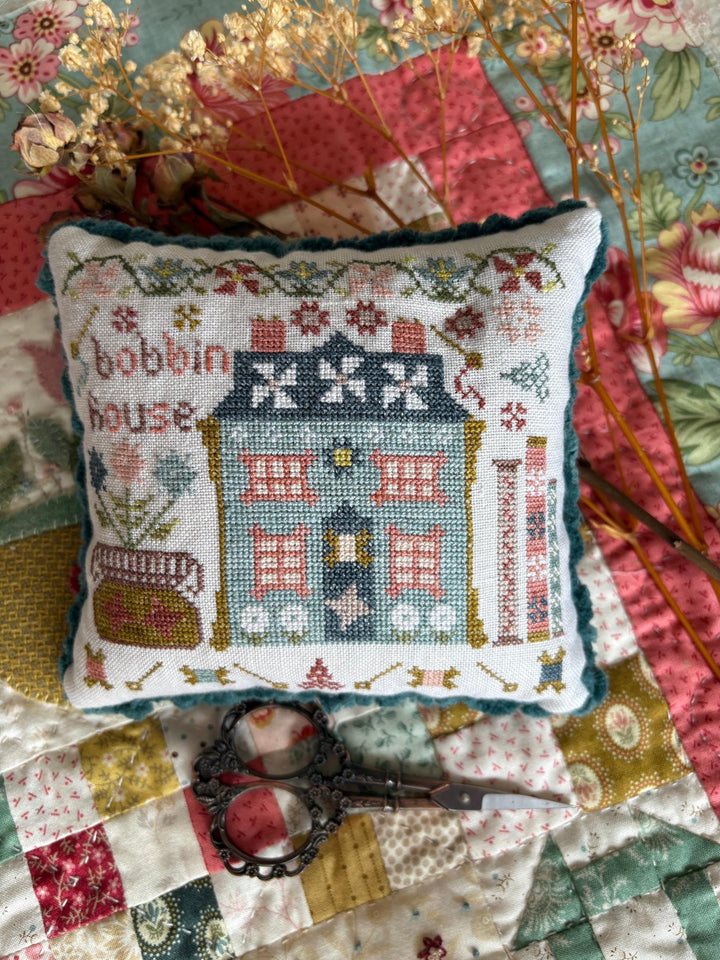 A square cross-stitch pillow featuring a colorful house design with the words 'bobbin house' on a patchwork quilt background, styled with embroidery scissors and dried flowers.
