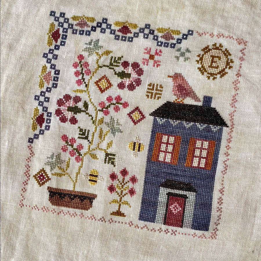 A cross-stitch needlework depicting a blue house with floral patterns and a birdcage.