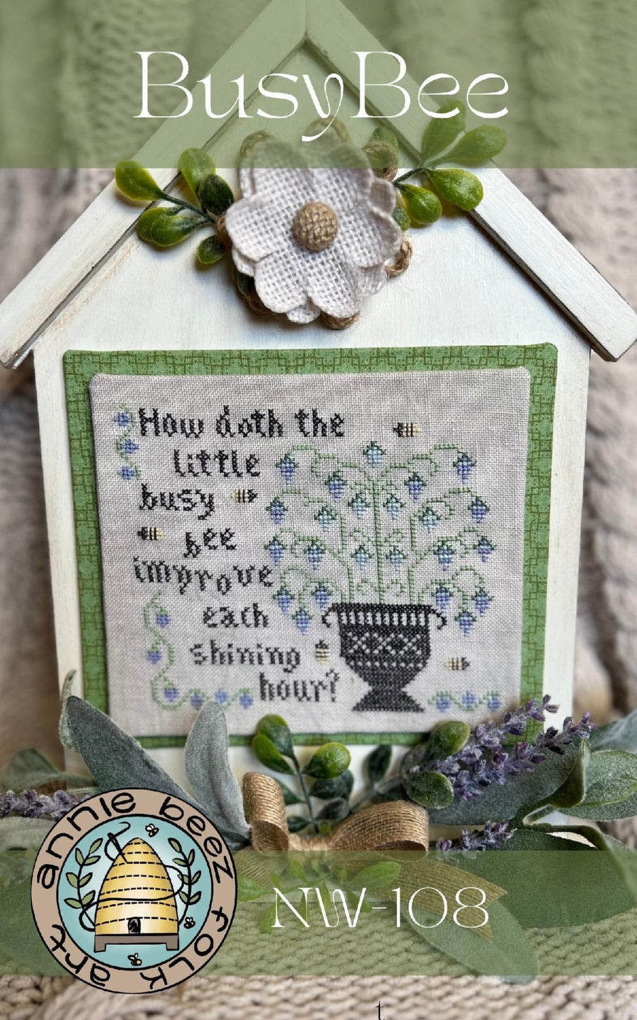 A stitched folk art piece with a green background, featuring a white text pattern that says 'How do the little bees Busy bees improve the shining hour?' surrounded by a green garden and a small house with a chimney.