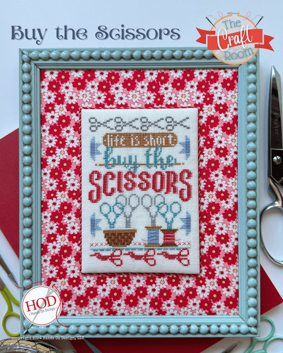 Framed cross-stitched design with 'Buy the Scissors' text on a floral background, with scissors and 'The Craft Room' branding.