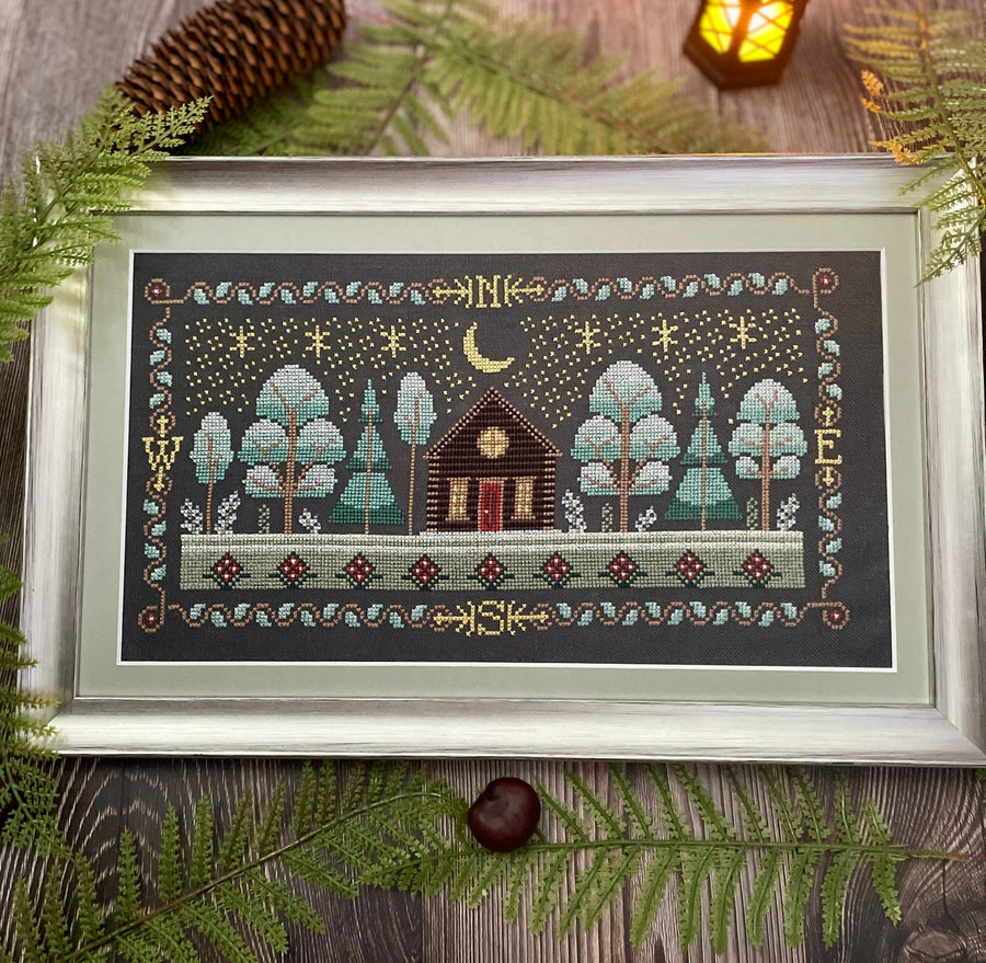 A framed needlepoint or cross-stitch artwork depicting a small cabin surrounded by trees under a crescent moon and stars, styled on a rustic wooden surface with pine branches and a warm lantern in the background.