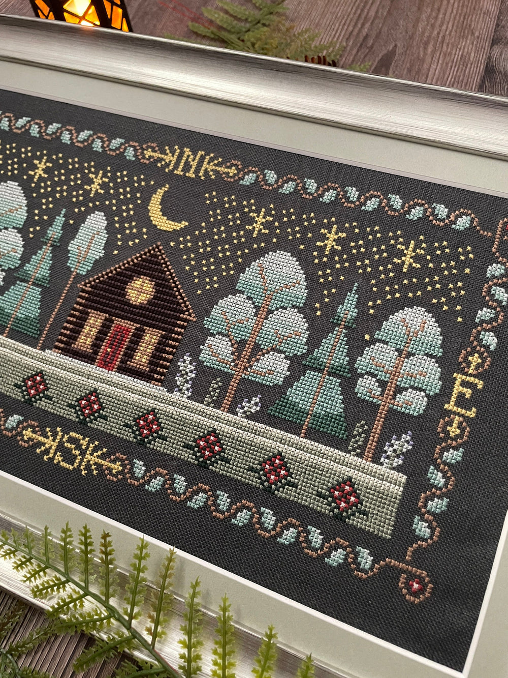 A framed needlepoint or cross-stitch artwork showing a cozy cabin, trees, stars, and a crescent moon on a dark background, photographed indoors with some plants and wood visible around it.