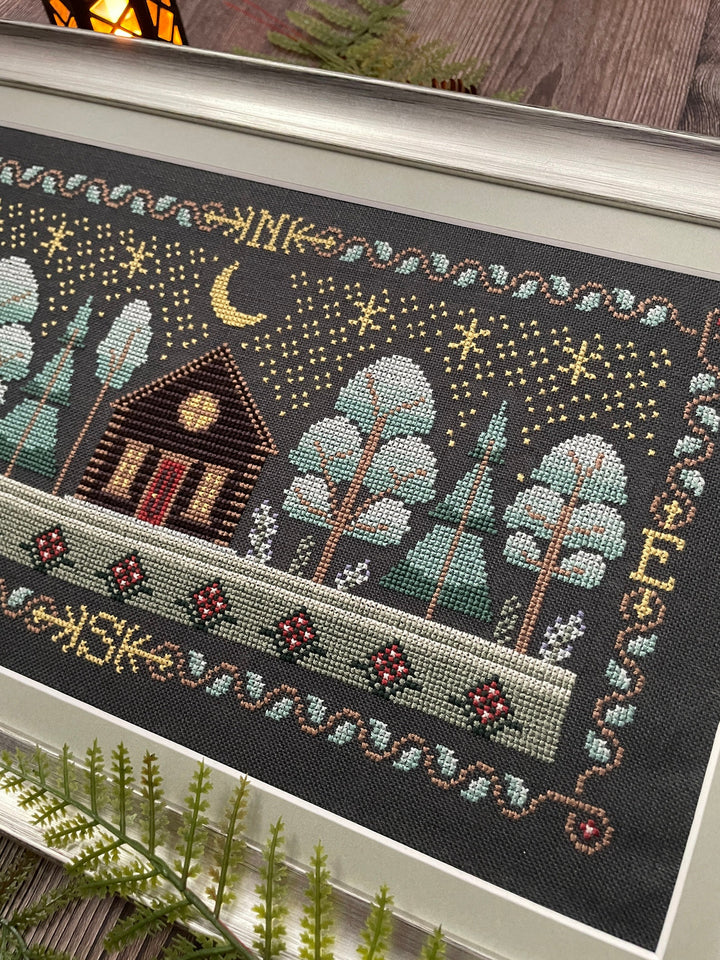 A framed needlepoint or cross-stitch artwork showing a cozy cabin, trees, stars, and a crescent moon on a dark background, photographed indoors with some plants and wood visible around it.