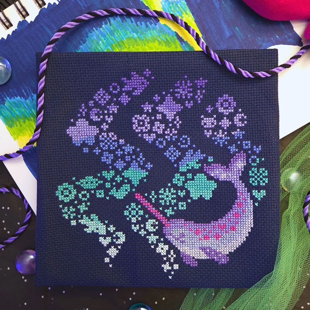 A square canvas cross-stitch design featuring a stylized mermaid in shades of purple, blue, and teal on a dark navy background, surrounded by colorful craft materials and a purple-and-white braided cord.