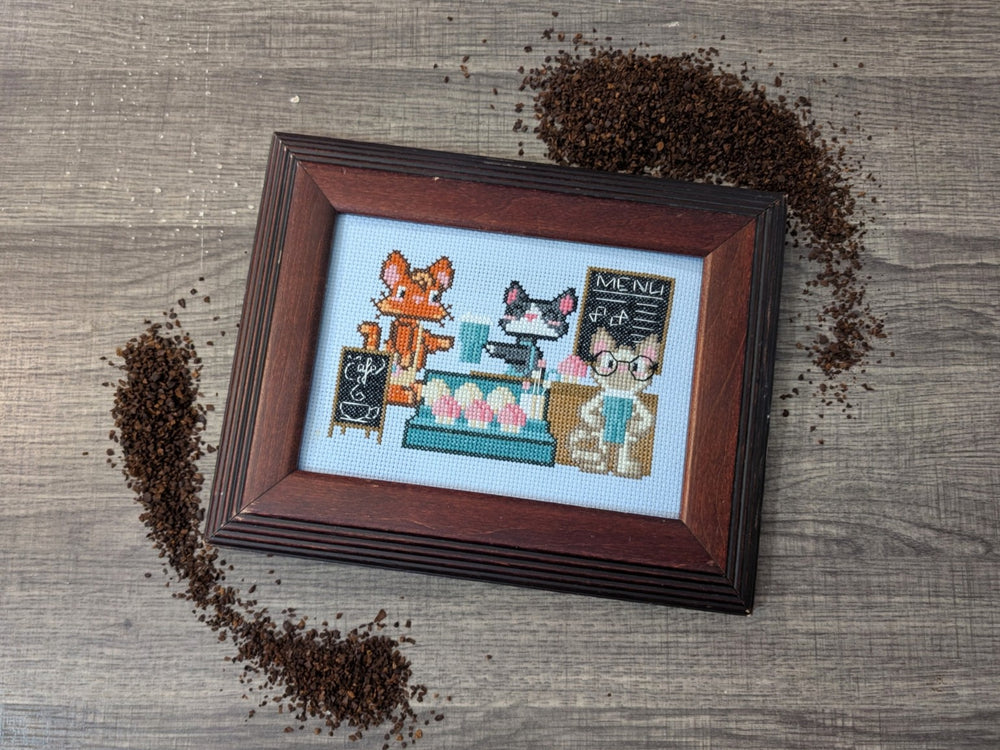 A framed cross-stitch artwork depicting a cozy cafe scene with a fox, a cat, and a person, styled on a gray wood-textured background with scattered coffee beans.
